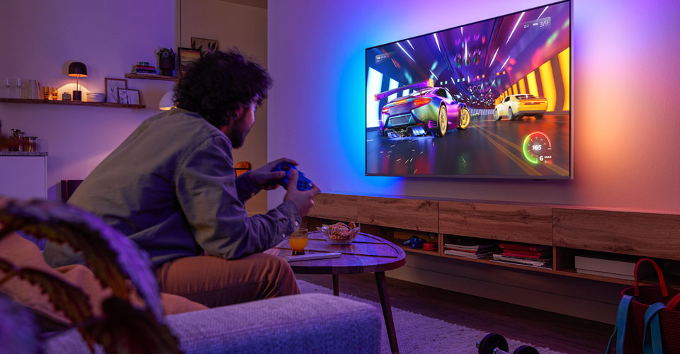 Žmogus sėdi ant sofos ir žaidžia lenktynių vaizdo žaidimą &bdquo;Philips Ambilight&ldquo; televizoriuje