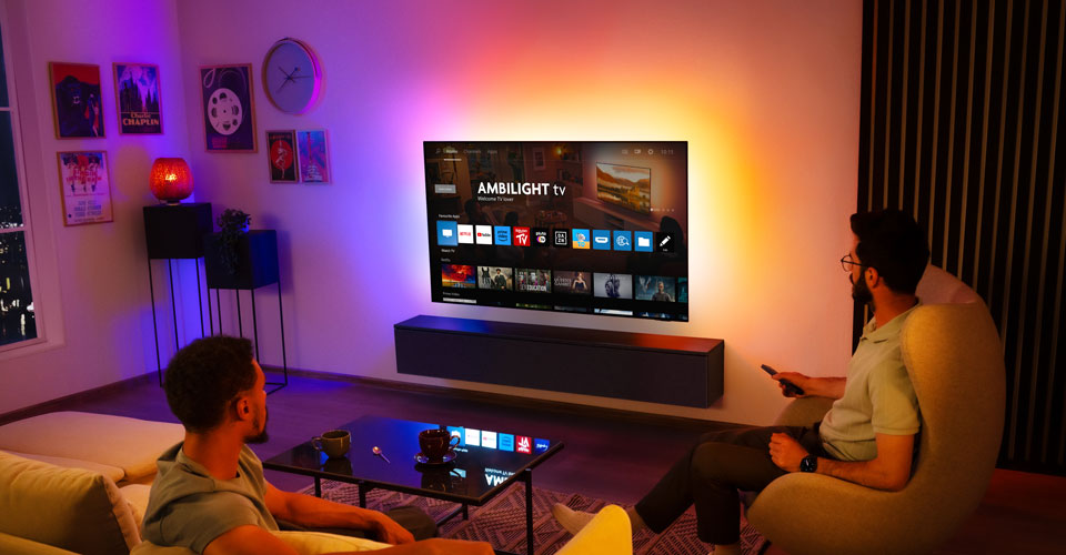 Du vyrai žiūri į prie sienos pritvirtintą &bdquo;Ambilight&ldquo; televizorių svetainėje